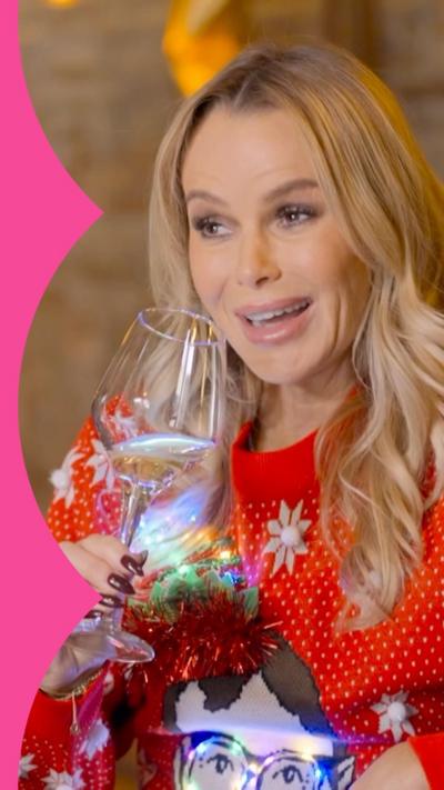 A woman, Amanda Holden, smiles while holding a wine glass. She has long blonde hair and wears a red festive jumper