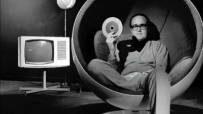 James Burke sits in a chair beside a tv, and holds tape for an electronic video recorder