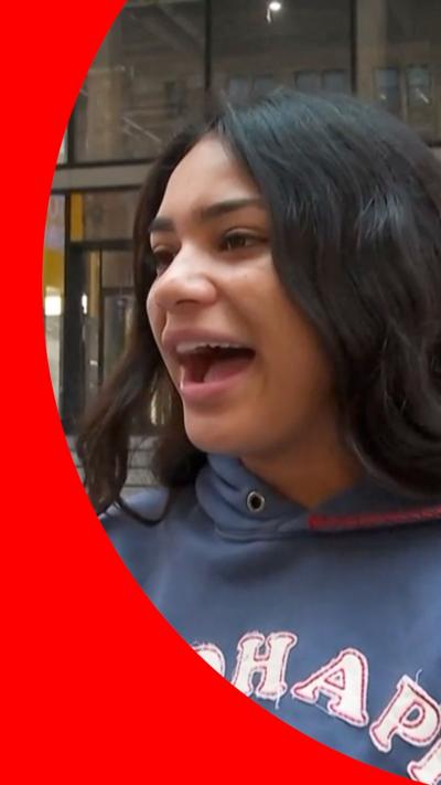 A woman with long wavy dark hair is talking excitedly. She is wearing a blue hoodie with letters appliqued to the front.