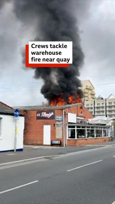 Black smoke and fire coming out the top of a brick warehouse building. 