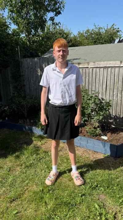 George is a schoolboy he is standing in a garden wearing a white school shirt and a black skirt. 