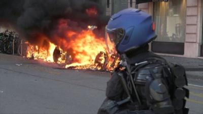 Police officer wearing protective gear watches car in flames