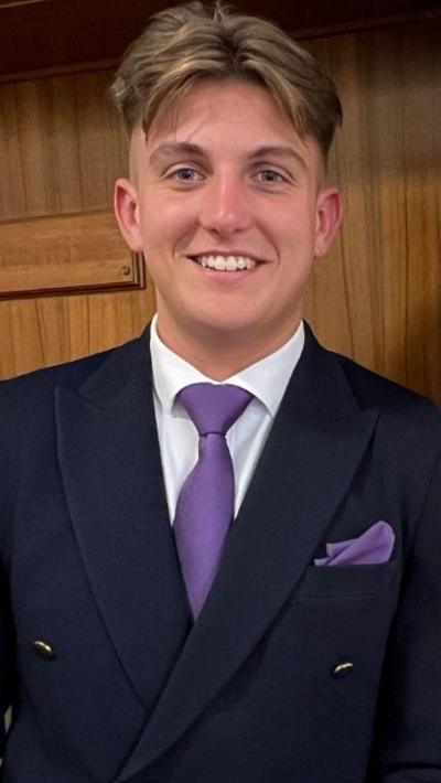 A man with brown hair with a centre parting in a blue suit with white shirt and purple tie