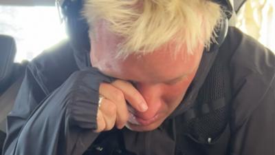 Jamie Laing, wearing headphones and sports gear, wiping tears off his face as he sits in a van