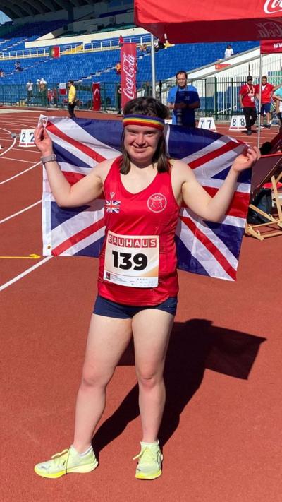 A woman standing on an atheltics track holding aloft a union jack. She's wearing a rainbow sweatband on her forehead, blue shorts and a red vest. Smiling at the camera.