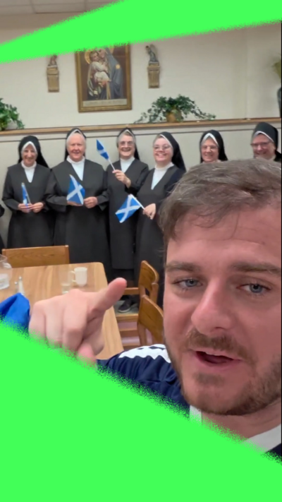 A man with brown hair and blue eyes with a short beard and moustache points at the camera. He is holding a Scotland flag and wearing a blue and white top. Behind him are nuns dressed in black and white and holding Scotland flags. They are standing in front of a wall with a religious painting hanging up beside plants.