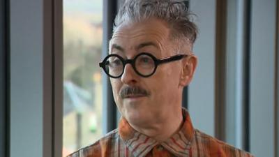 Actor Alan Cumming in front of a grey background. He has grey, sticking up hair; thick, black rimmed round glasses and a moustache. He is wearing an orange and grey checked shit.