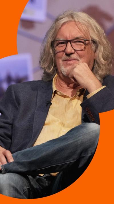 James May has long wavy grey hair and glasses. He is wearing jeans, a yellow shirt and a navy blazer.