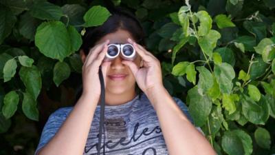 A young teenager looks through binoculars from a bush