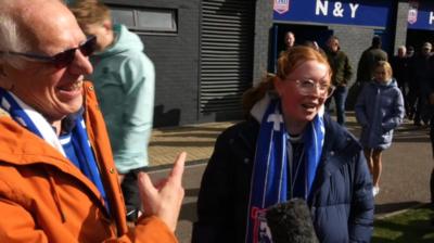An older man in an orange jacket and Ipswich Town's blue underneath, stands facing a reporter who is off camera. He is pointing to a young woman with red hair beside him, who is in a navy coat.