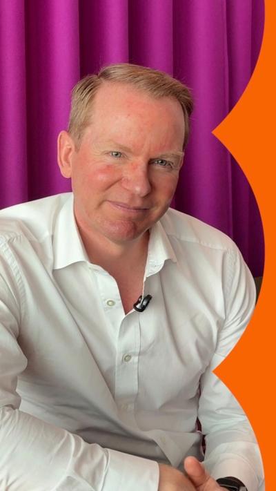 A man, Charlie Nunn, wears a white shirt. He has short red hair and sits in front of a purple curtain.