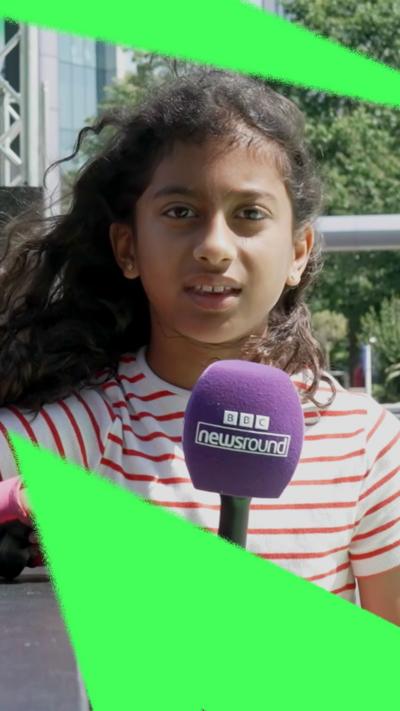 A young girl standing on a football pitch, holding a BBC Newsround microphone