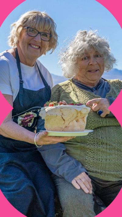 Baker Angie Taylor holding a pavlova while Miriam sits next to her and points to it