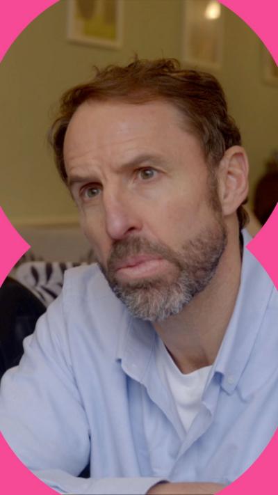Gareth Southgate has his brown hair, a beard and moustache. He is wearing a white vest and a pale blue shirt.