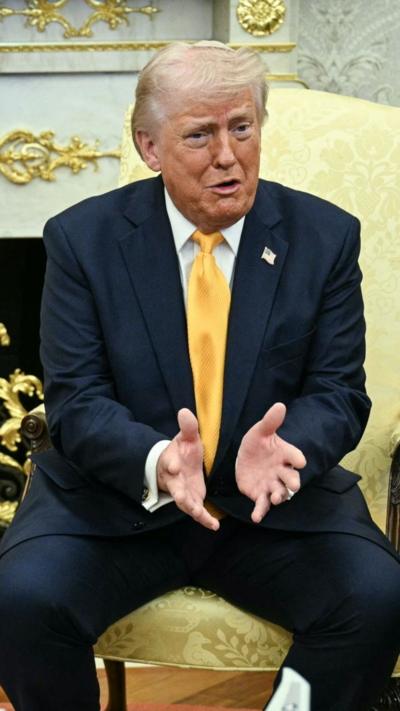 Donald Trump sits in the Oval office gesturing with his hands.