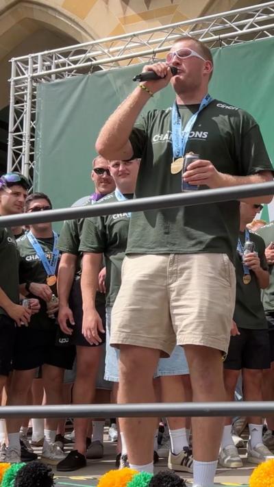 A man wearing a green sports jersey and a medal is holding a microphone. His team mates are stood behind him in front of a large green stage background