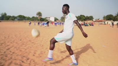 Yankuba Minteh in light blue kit faces left and juggles a football on a sand pitch