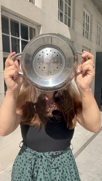 A woman holds a colander over her face