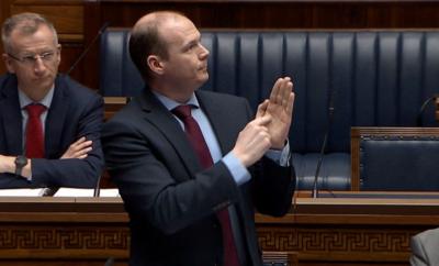 Lyons is standing in the chamber and is wearing a navy suit with a red tie. His hands are raised as he is using sign language.