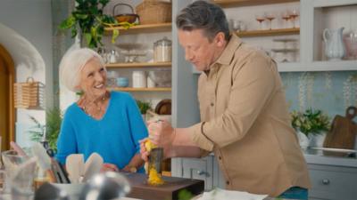 Mary Berry smiles at Jamie Oliver in a kitchen. He is grating a lemon on a chopping board