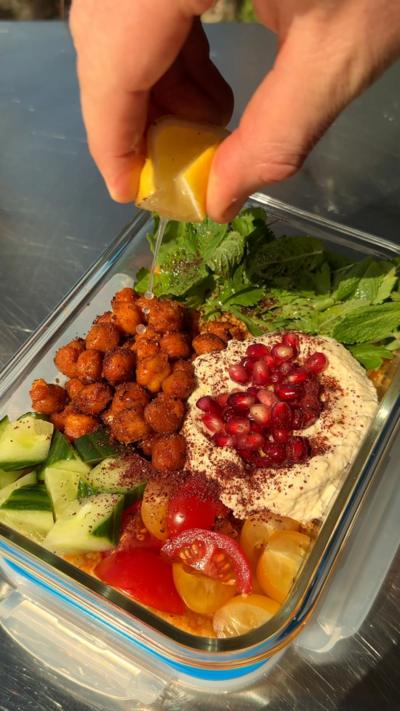 Harissa couscous with crispy chickpeas in a food storage container with a lemon being squeezed on top of it