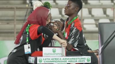 Two female arm wrestlers, one wearing a head scarf, clasp hands and hold handles on a table as a partially-obscured referee prepares them for a contest