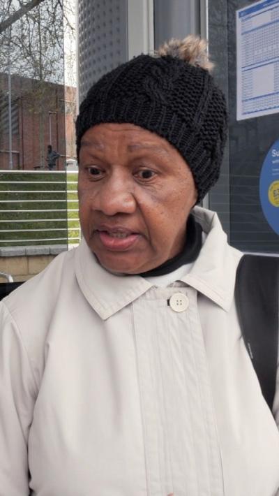A black woman in a white coat and black bobble hat talking at a tram stop