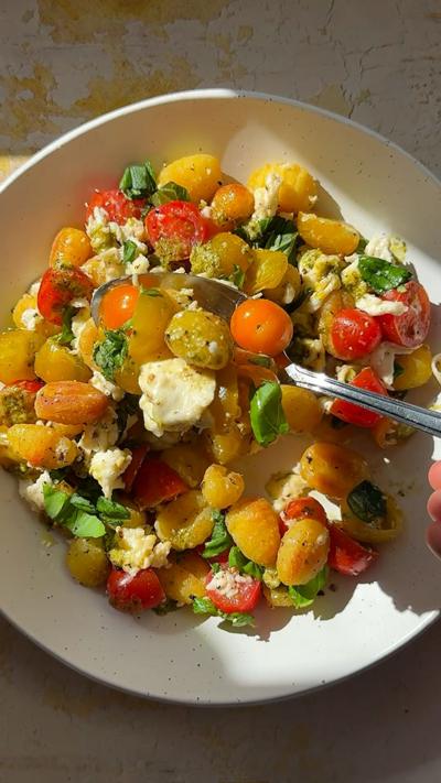 A white plate of roasted gnocchi with cherry tomatoes, basil and mozzarella cheese, with a spoon shown coming in from the side.
