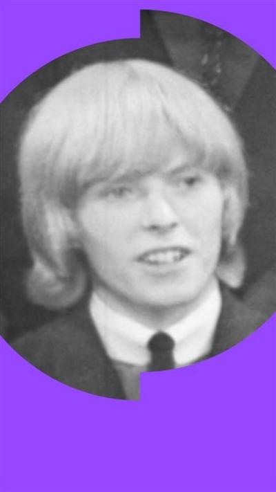 Black and white photo of David Bowie in 1964 wearing a suit and tie, with shoulder length hair and a fringe.