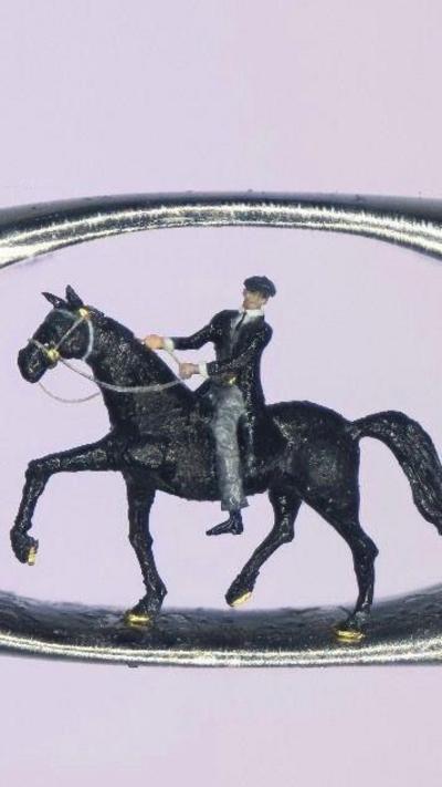 A micro sculpture of a man in a black coat, black hat and grey trousers sat in a horse inside the eye of a needle