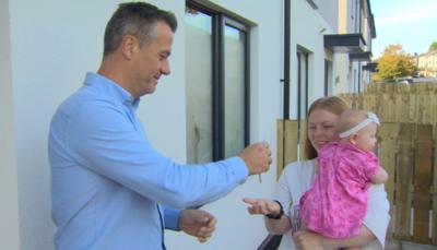 Man in blue shirt hands keys to ginger woman in white t-shirt holding a baby in a pink dress and white hairband 