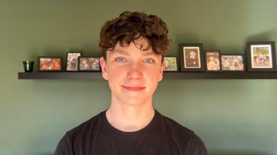 Fiachra, air a bheil dyslexia, na sheasamh air beulaibh chamera agus lèine dhubh air. 