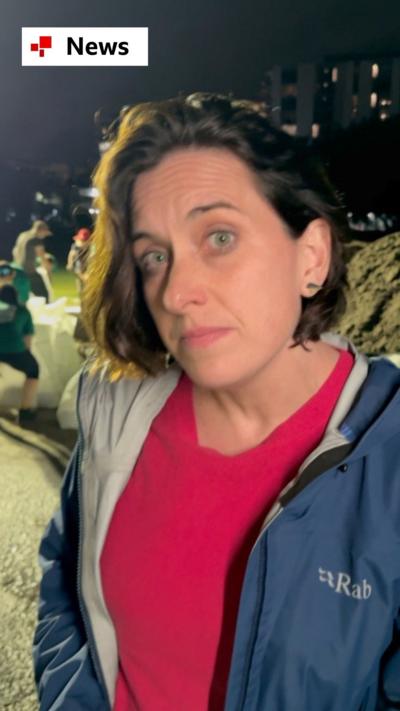 BBC reporter Katy Watson wears a red top and blue rain jacket. People filling sandbags can be seen behind her.