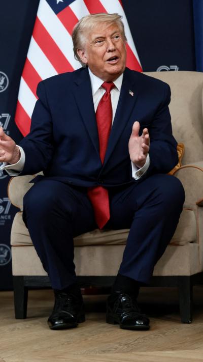 Donald Trump speaking. He is wearing a red tie and is sat in front of the US flag.