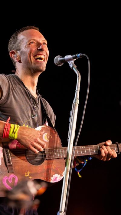 Chris Martin smiles while holding a guitar as Coldplay perform at Glastonbury 2024