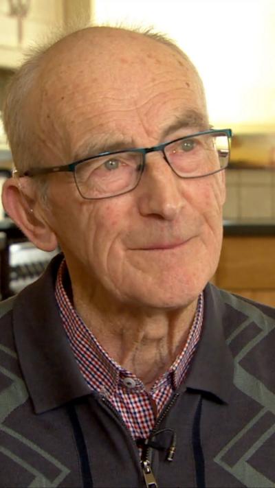 Alphie Lonergan is bald and is wearing a grey jumper with a shirt underneath.