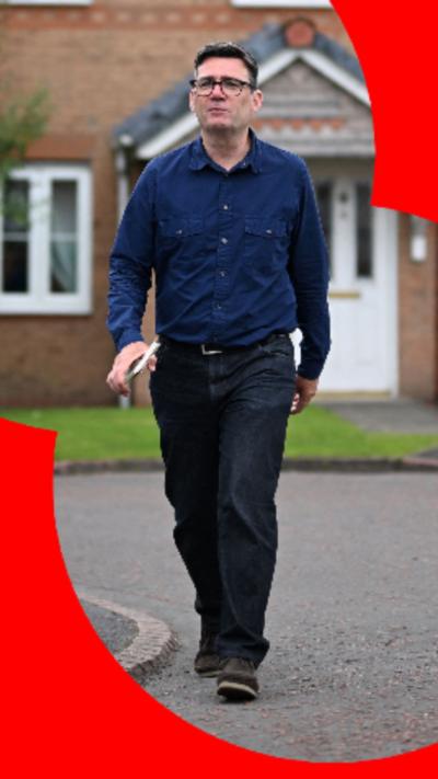 Andy Burnham wearing a dark blue shirt, walking away from a house with a white door.