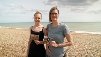 Two women pose holding postcards on Hastings beach
