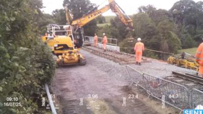 A yellow digger lowers new track on to a railway line. There are four engineers in orange uniforms supervising the work