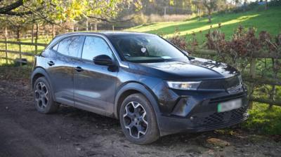A black SUV-type car is parked on a muddy track, next to a fence and a field