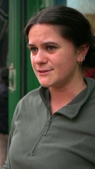 A woman looks slightly off camera. She has dark hair tied up and has a khaki top on. It is a head and shoulders shot