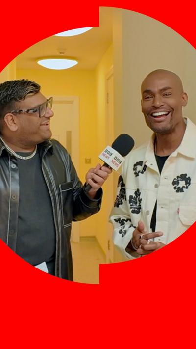 Paul C Brunson laughs while being interviewed by Jameel Shariff in a yellow corridor