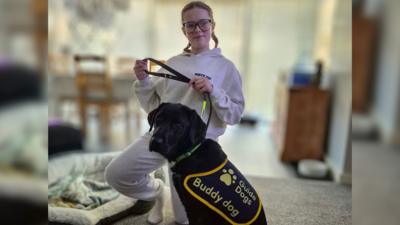 Aniyah holds onto Spirit's lead. She is a black lab wearing a Guide Dogs coat which says Buddy Dog. Aniyah is smiling