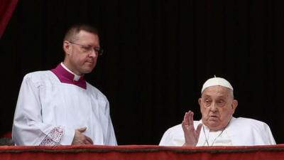Pope Francis sits and waves from a chair on a balcony next to a colleague.