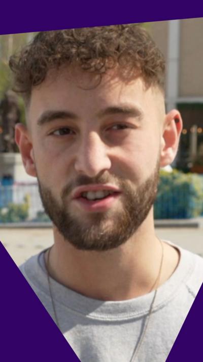 A young man with brown hair and beard in a grey top and chain around his neck.
