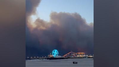 Plumes of dark grey smoke are seen above a lit-up coastal theme park on Santa Monica's iconic skyline 