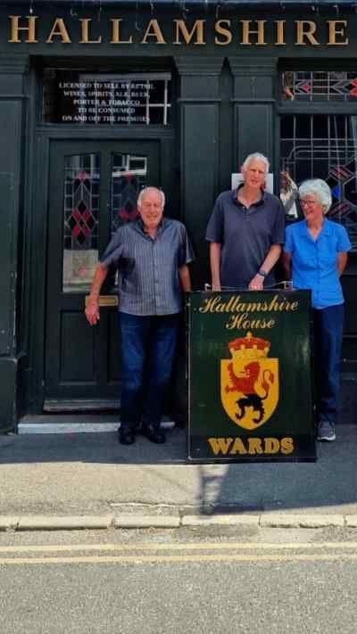 People stood outside a pub. They are holding a pub sign. 