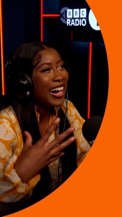 A woman in a radio studio smiles with one hand raised.