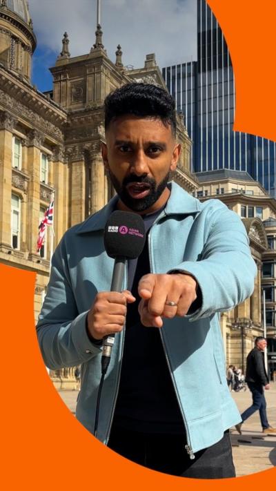 A man in a blue jacket holds a microphone in one hand and points at the camera with the other