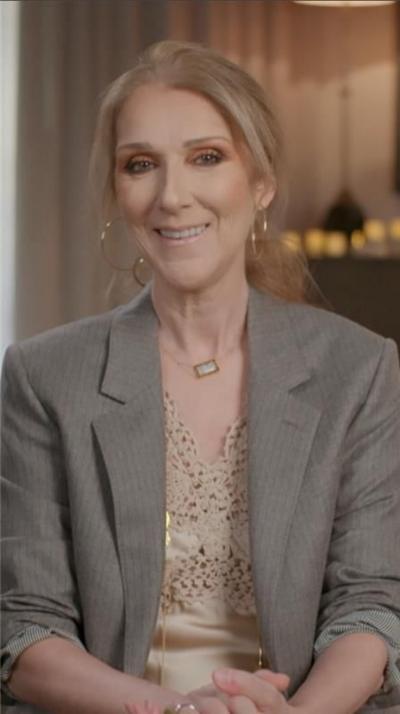 Céline Dion sitting on a chair with flowers behind her, smiling and wearing a grey blazer with a cream top